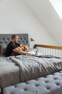 Side view of male sitting on bed with glass of orange juice and browsing tablet while having breakfast in morning at home