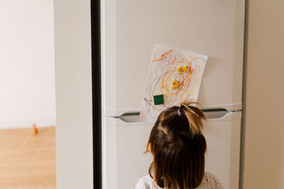 Rear view of girl looking at drawing on refrigerator in home