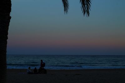 Silhouette people sitting on beach against clear sky during sunset