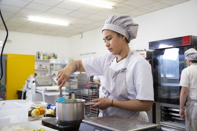 Female cook in uniform stirring ingredients in saucepan while preparing syrup on stove in professional light bakery school