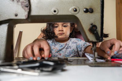 Unrecognizable seamstress with cute hispanic girl sewing on professional rusty machine while sitting at table with supply in light workshop