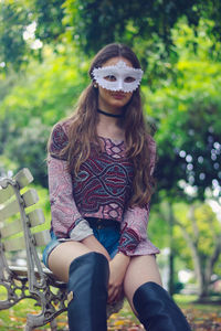 Portrait of smiling young woman wearing venetian mask while sitting on bench at park