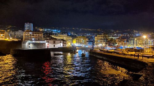 Illuminated buildings by sea against sky at night