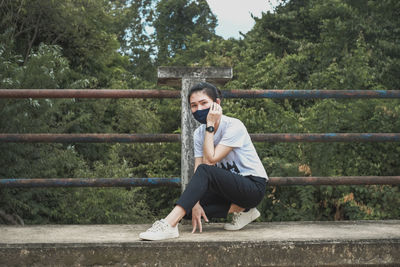 Portrait of young man sitting on fence against trees