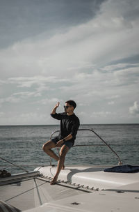 Full length of man shielding eyes while sitting in boat on sea against sky