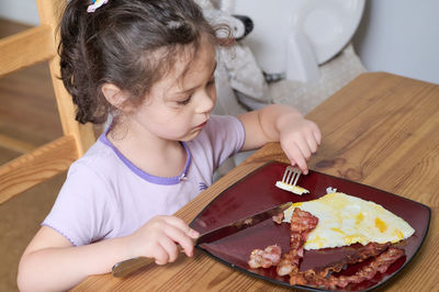 Young girl eating eggs and bacon with knife and fork for the first time