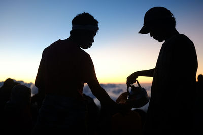 Silhouette man pouring coffee for hiker against sky during sunset