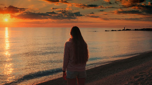 Rear view of woman standing at beach against sky during sunset