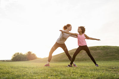 Mother assisting daughter practicing exercise on grass at park against sky