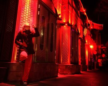 Full length of man holding cigarette by illuminated building on sidewalk in city at night