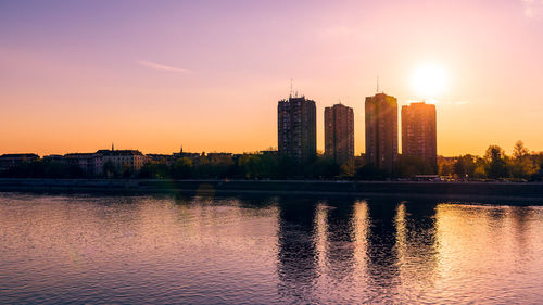 Modern buildings by river against sky during sunset