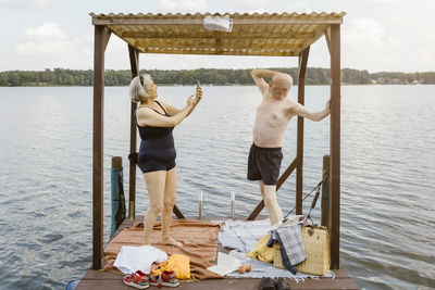 Full length of senior woman photographing shirtless man while standing in gazebo against river