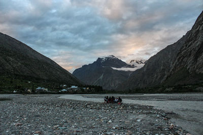 People at riverbank by mountains against cloudy sky