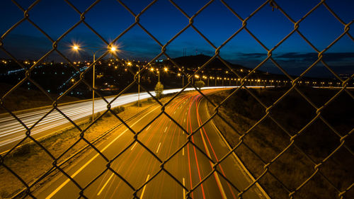 High angle view of light trails on highway seen through chainlink fence at night