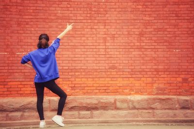 Rear view of woman with hand raised posing against brick wall