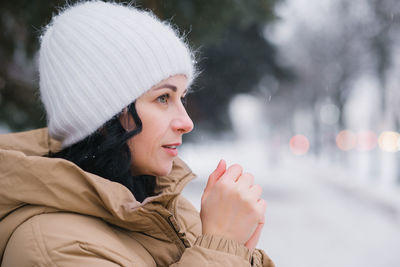 A happy woman stands in a coat and warms her hands. it stands in the city in winter under the snow