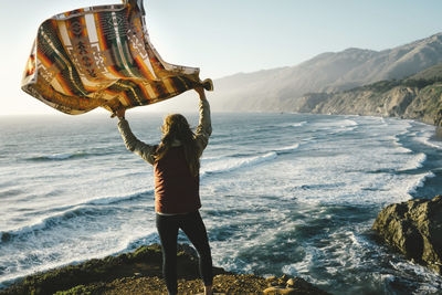 Rear view of woman holding scarf while standing on rock against sea