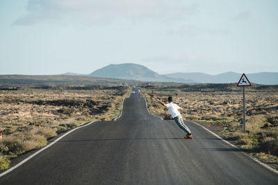 Full length of young man skateboarding on country road against sky