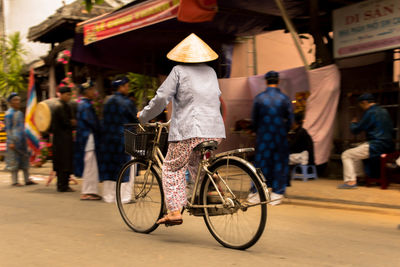 Rear view of woman wearing conical hat cycling on street in city