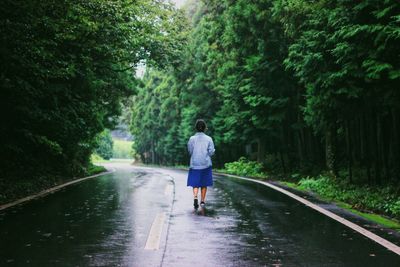 Rear view of woman walking on wet road amidst trees during rainy season