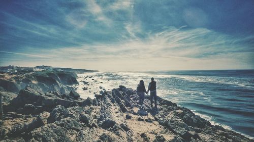 Rear view of couple standing in cliff by sea against sky