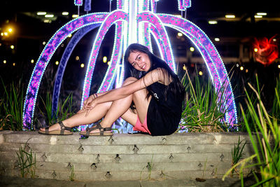 Full length portrait of smiling young woman sitting by illuminated fountain at night