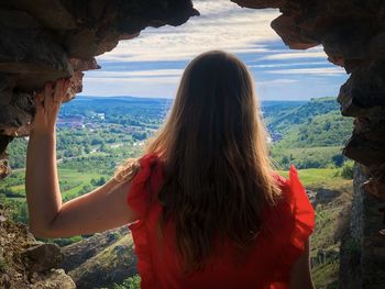 Woman in red standing near an opening in a stone wall and looking at the panorama in front of her