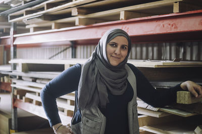 Portrait of smiling female carpenter standing with hand on hip by pallet stack in warehouse