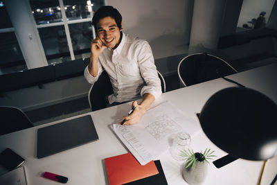 High angle portrait of smiling businessman sitting with blueprint at desk while working late in office