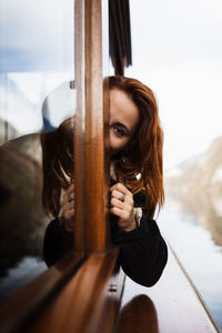 Young female tourist in warm wear looking out open window while travelling by boat on calm lake surrounded by rocky mountains in autumn day