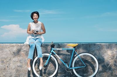 Portrait of young woman standing on bicycle against sky