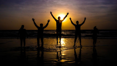 Silhouette friends with arms raised on shore at beach during sunset