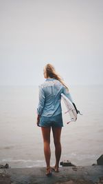 Rear view of young woman with surfboard standing at beach against sky