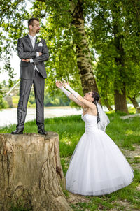 Side view of bridegroom reaching for bride standing on tree stump at park