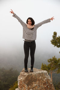 Full length of carefree young woman with arms raised standing on rock during foggy weather