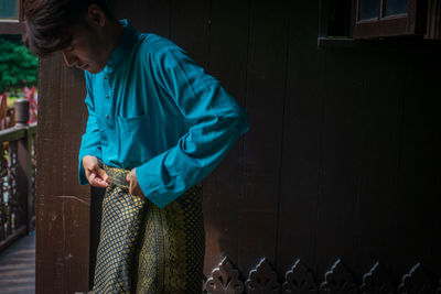 Side view of young man wearing sampin in traditional clothing in a malay wooden house during eid. 