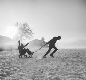 Brother pulling sister on sled at snow covered field against sky