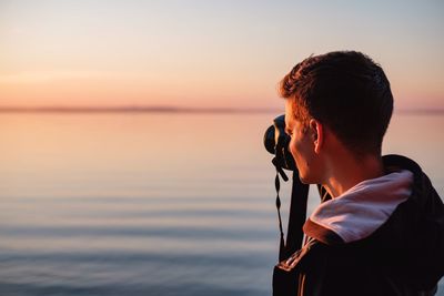 Young man photographing sea against sky during sunset