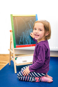 Side view portrait of cheerful girl chalk drawing on blackboard at home