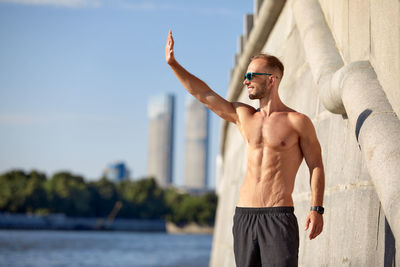 Rear view of shirtless man with arms raised against lake