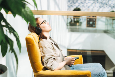 Young teenager girl student using mobile, listen music in headphones in yellow chair at public place