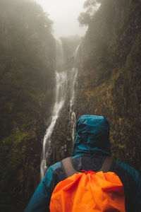 Rainy weather hiker in a waterproof jacket looks at the majestic risco waterfall immersed in mist 