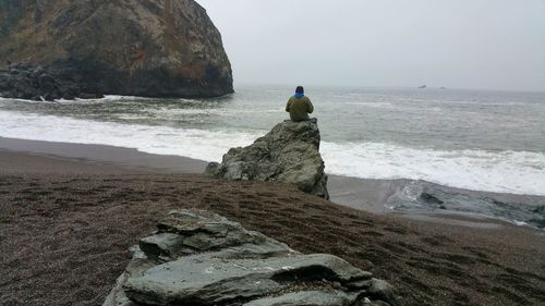 Rock formation with man sitting on boulder gazing out to sea on beach against sky