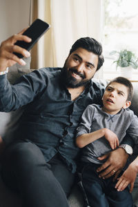 Smiling father taking selfie with autistic son while sitting on sofa in living room