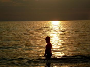 Silhouette boy in sea against sky during sunset