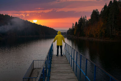 Rear view of man walking on pier over lake against sky during sunset