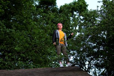 Low angle view of young man standing on rooftop against trees