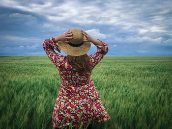 Rear view of young woman with a hat standing in a wheat field on a day with clouds