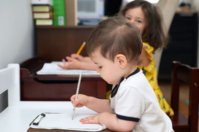 Little toddler copying his sister doing her homework