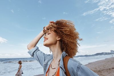 Portrait of young woman standing at beach against sky